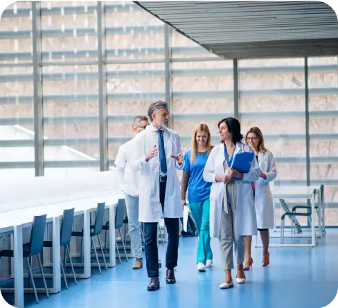 Grupka lekarzy w białych fartuchach i pielęgniarka w niebieskim uniformie idą przez korytarz i rozmawiają – nawiązanie do branży medycznej.