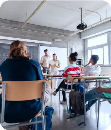 Lekcja w klasie, przy tablicy stoi nauczyciel oraz uczennica z kartką papieru w dłoniach, przy ławkach siedzą uczniowie.