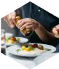 Dwa talerze z jedzeniem na stole, a przy nim mężczyzna, który trzyma w dłoniach młynek do przypraw – nawiązanie do gastronomii.
